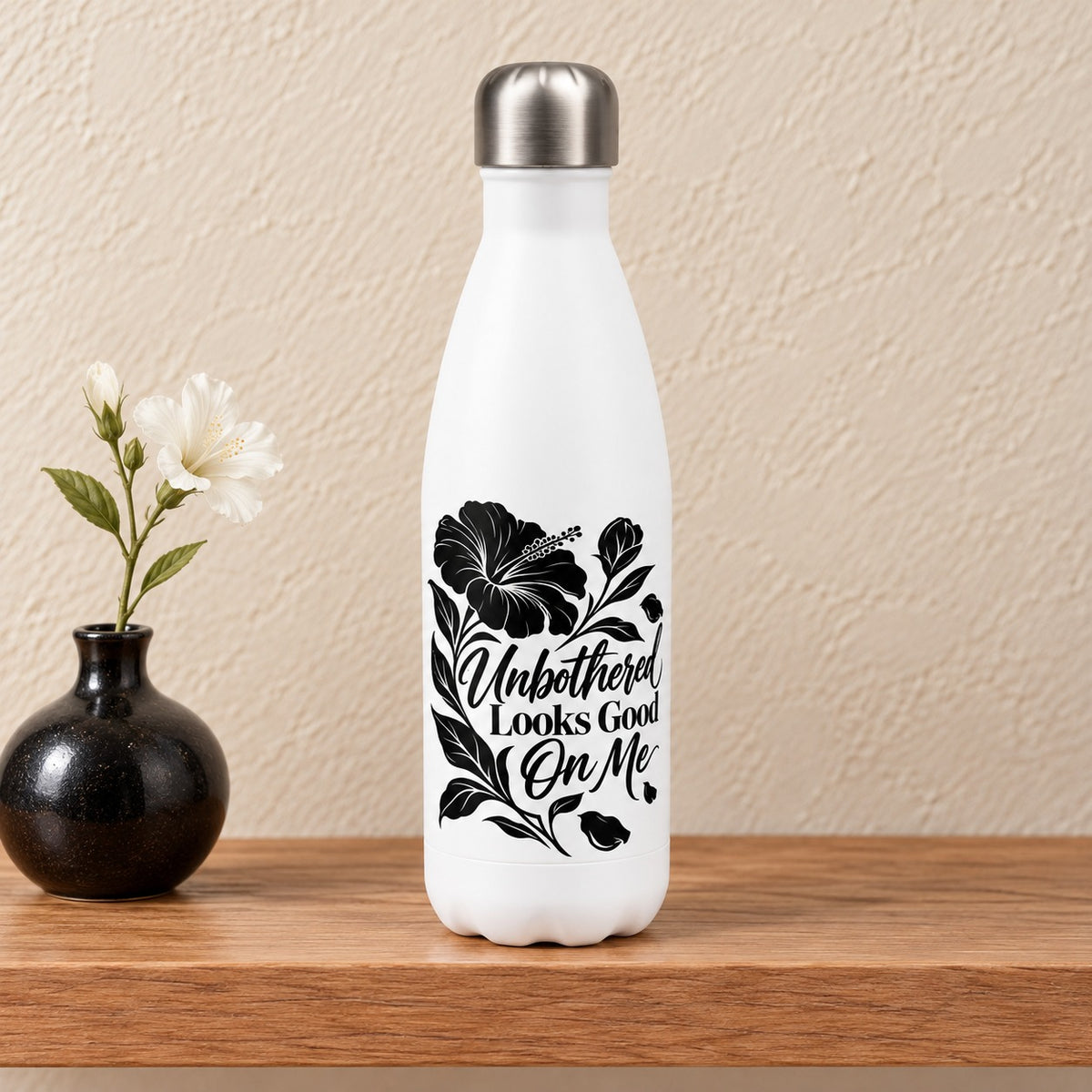 Unbothered Looks Good On Me 17 oz white water bottle with a bold black hibiscus and script lettering, standing on a light oak shelf beside a black bud vase