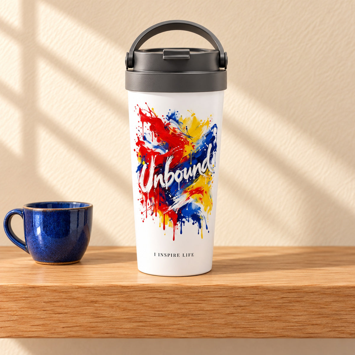 Unbound 15 oz white stainless steel travel mug on a light oak shelf beside a small cobalt blue ceramic cup, design facing forward