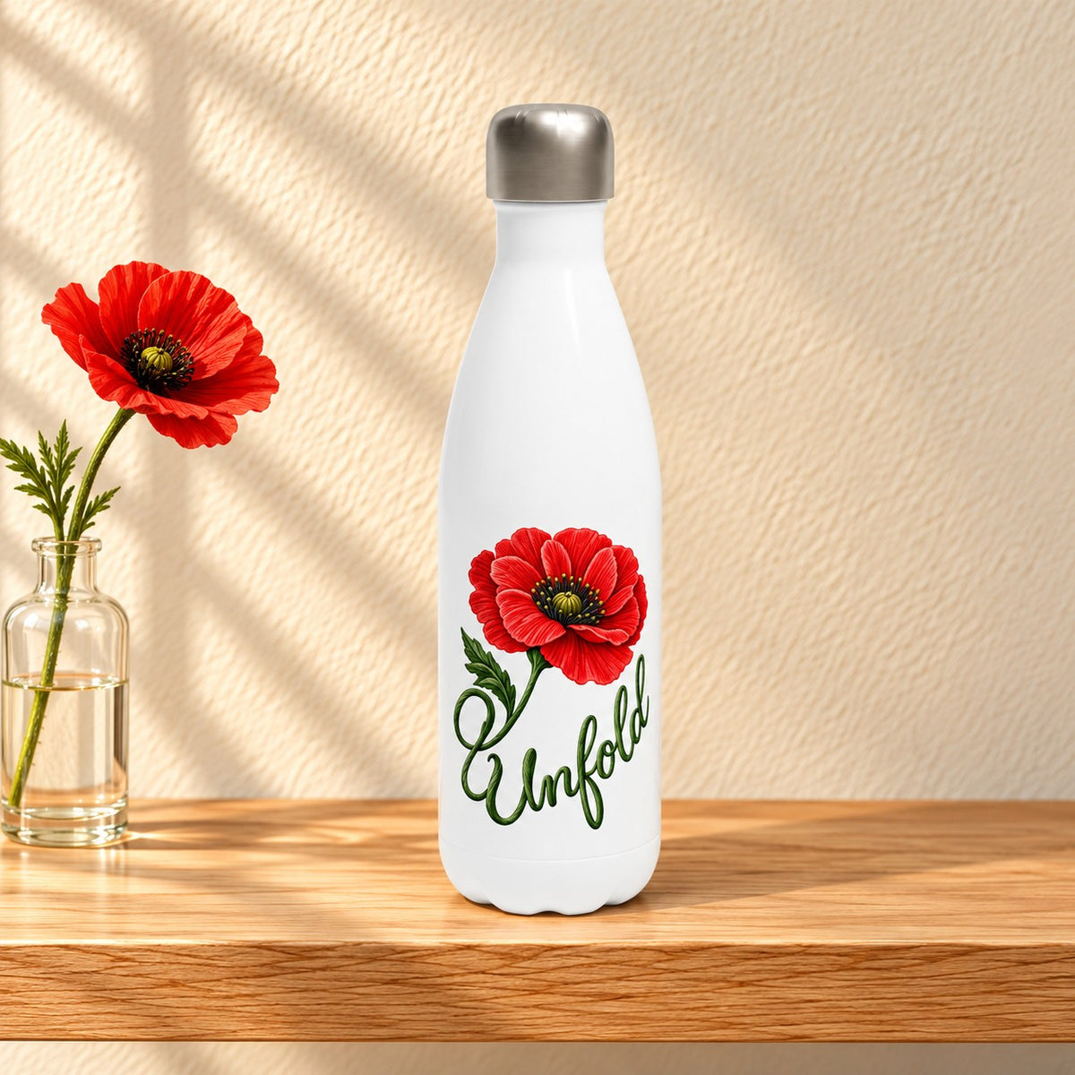 Unfold white 17 oz water bottle with a red poppy whose stem curls into the word Unfold, standing on a light oak shelf beside a poppy in a glass bottle