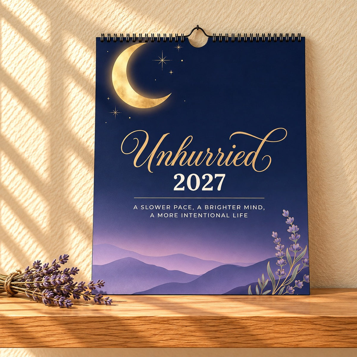 Unhurried 2027 wall calendar with a gold crescent moon over lavender hills, standing on a light oak shelf beside dried lavender