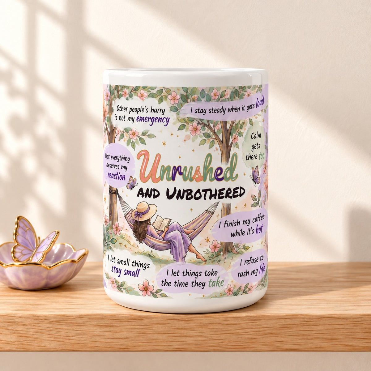 Unrushed and Unbothered 15 oz white mug with the title facing forward, resting on a light oak shelf beside a lilac butterfly dish