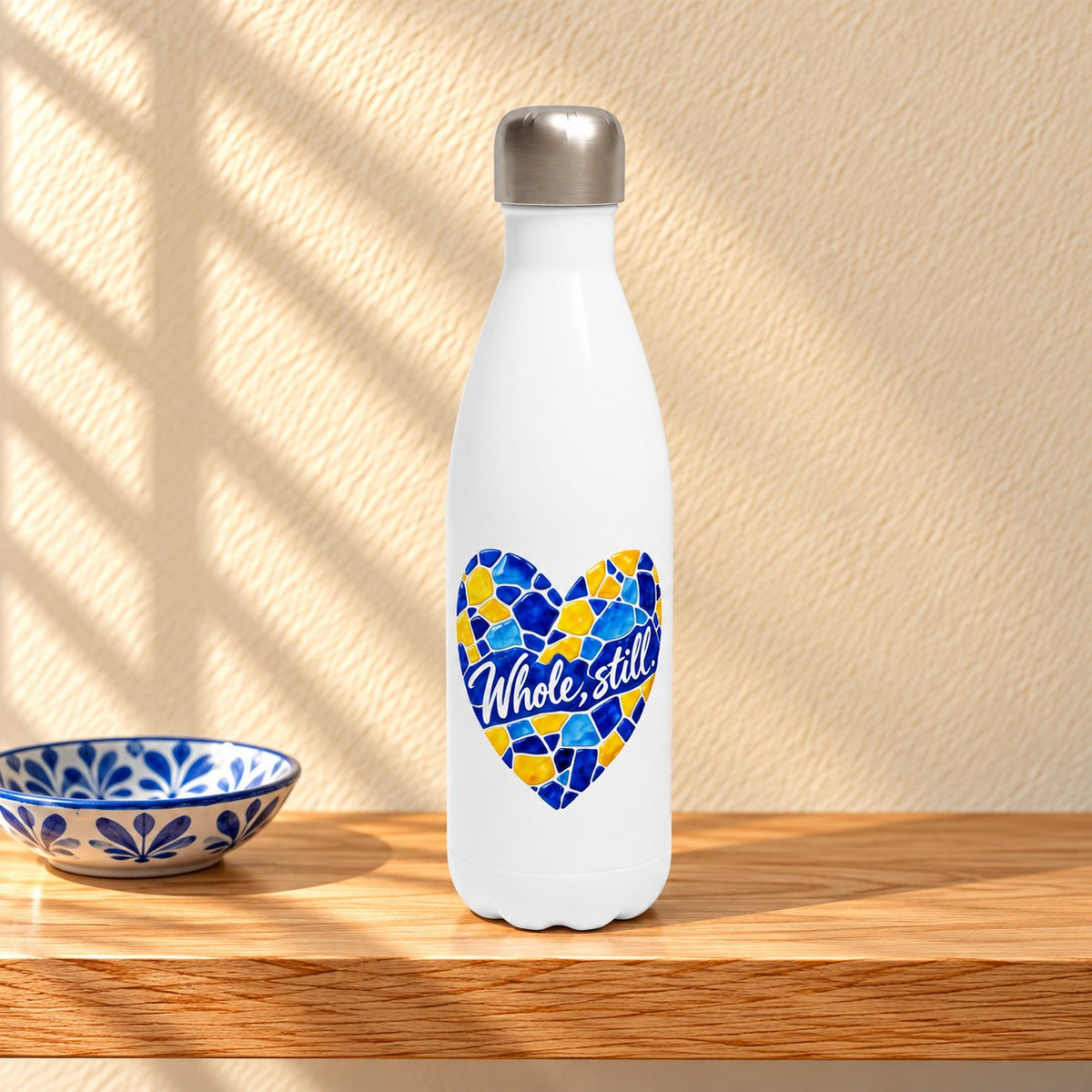 Whole, Still white 17 oz water bottle with a blue and yellow mosaic heart, standing on a light oak shelf beside a blue and white ceramic dish