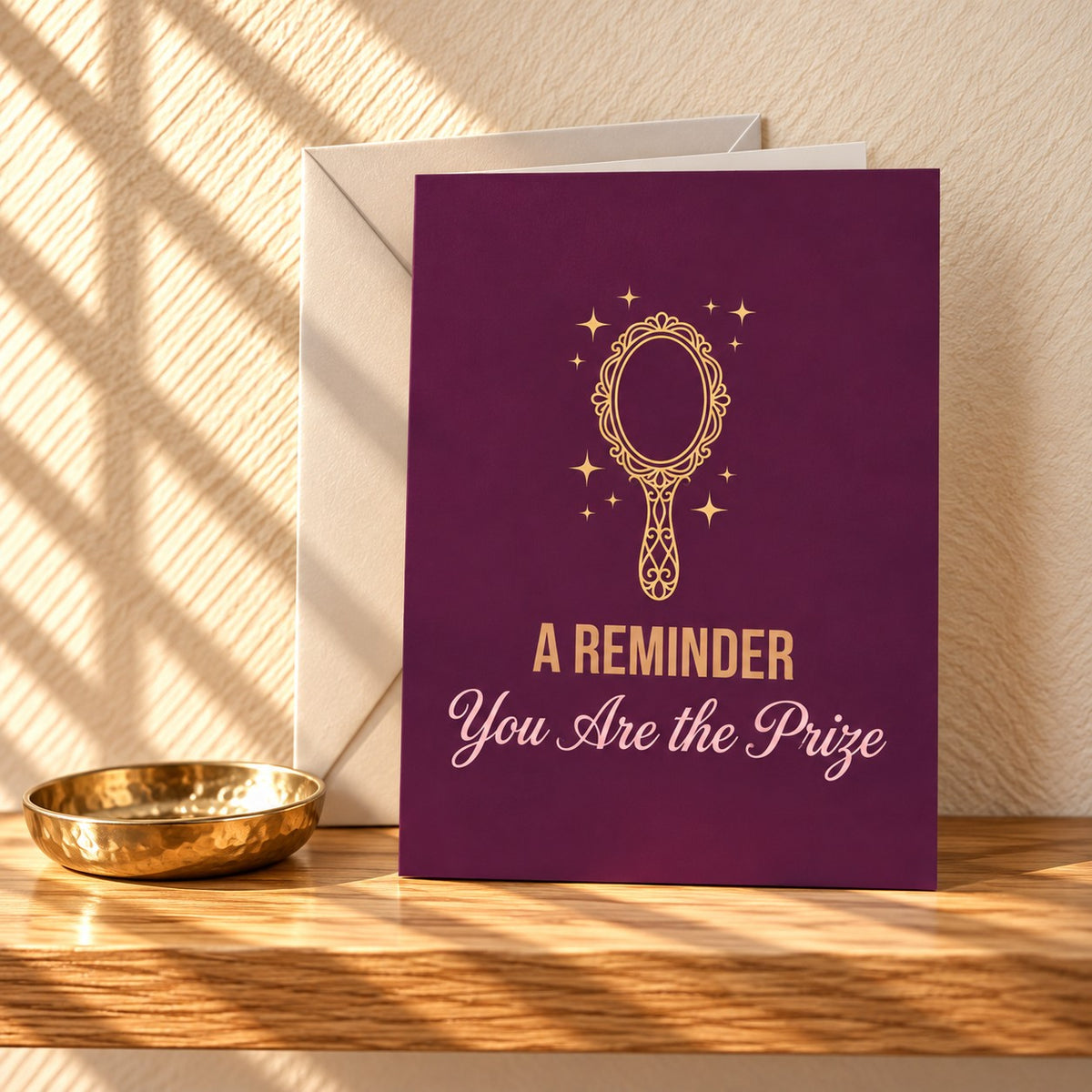 A Reminder You Are the Prize card in deep plum with a gold hand mirror, standing on a light oak shelf beside a gold trinket dish